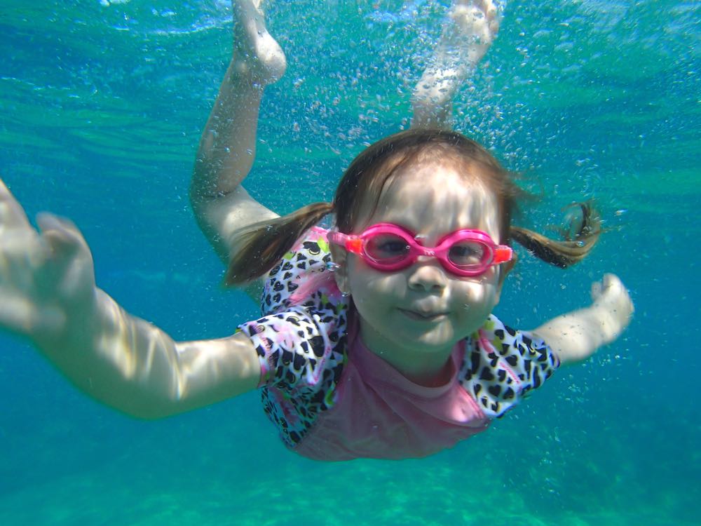 An underwater photo of a 3 year old girl wearing pink goggles swimming in the clear blue maui ocean.